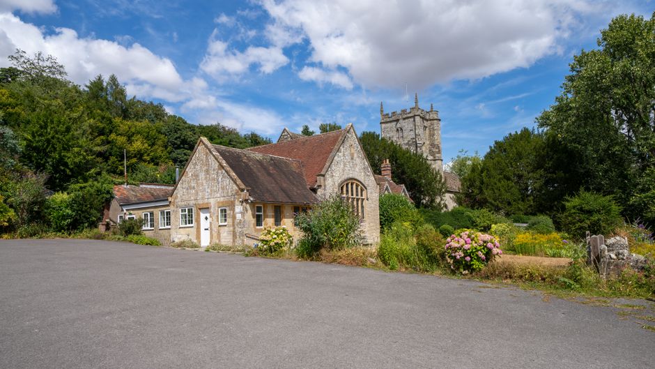 A scenic view of a rustic stone cottage with a sloped red-tiled roof, situated in a garden setting on Hall Road, Wennington. The garden features various plants, including flowering bushes with pink bl